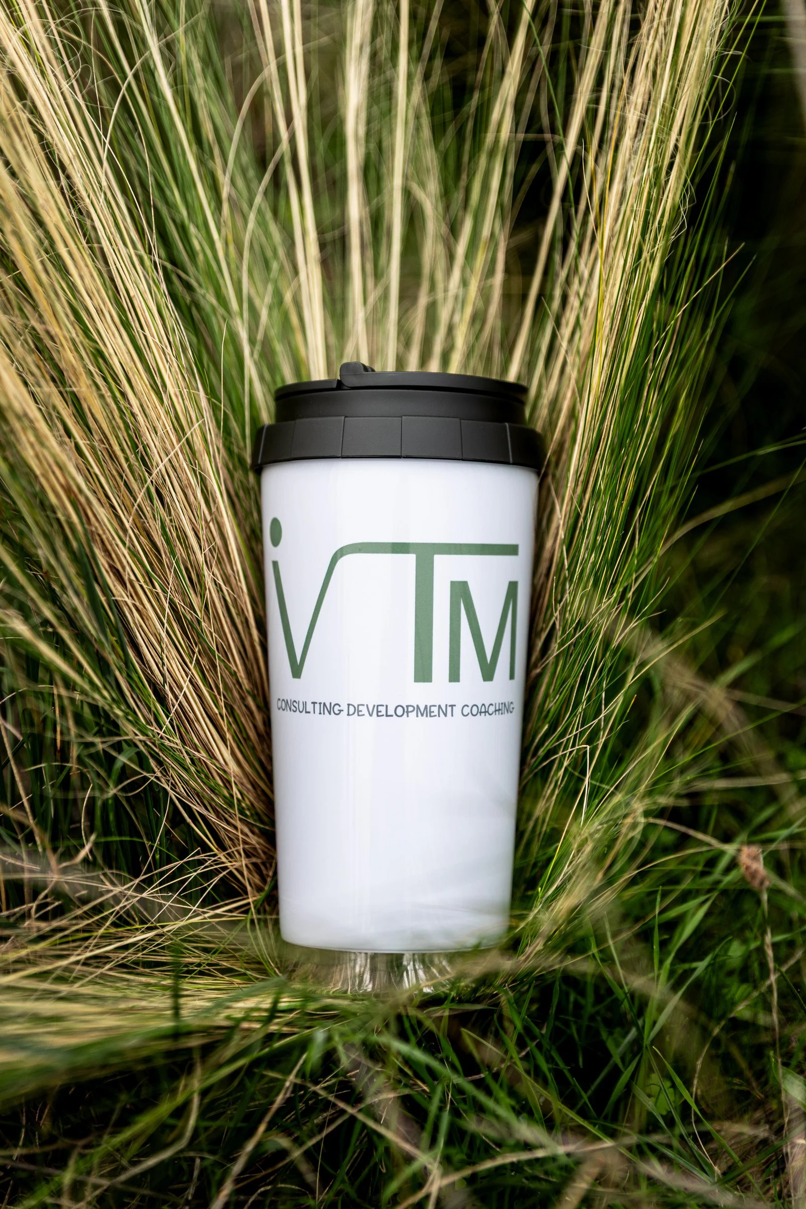 Ein weißer Becher mit dem IVTM-Logo steht zwischen hohen Gräsern